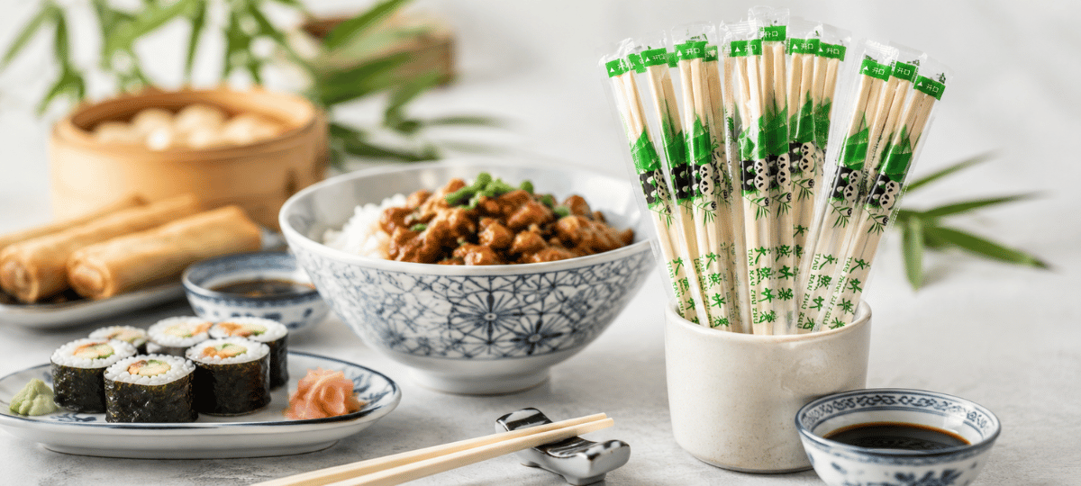 Einweg-Essstäbchen aus Bambus in hygienischer Einzelverpackung, stilvoll präsentiert in einer asiatischen Tischdekoration mit Sushi und weiteren Speisen.